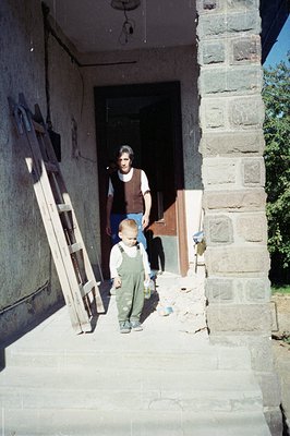 A young boy in overalls stands on a stone-paved porch, carrying a bottle. An adult, presumably a caretaker, stands in the doo...