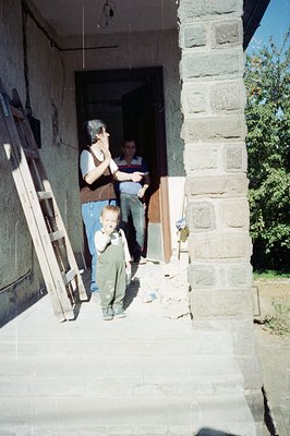 A young boy in overalls stands on a stone porch. Two men stand inside an open doorway, seemingly observing construction work....