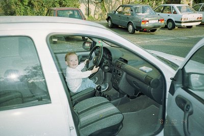 A young child, appearing to be a toddler, sits in the driver’s seat of a white sedan, mimicking driving with both hands on th...
