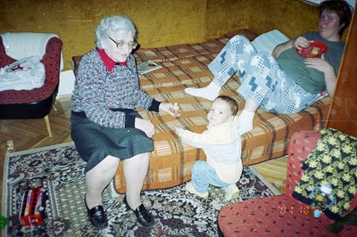 An elderly woman in a patterned dress and black shoes sits with a toddler reaching towards her. A young woman in casual attir...