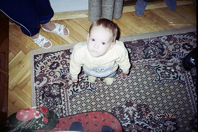 Infant wearing a cream-colored sweater stands on a patterned area rug with floral and geometric elements. The photograph’s pe...