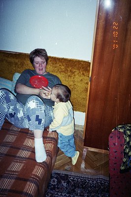 A young boy in overalls playfully reaches toward a woman seated on a patterned sofa. She's wearing a patterned pajama top and...
