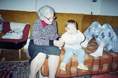 An elderly woman with a red sweater and plaid skirt sits on a floral sofa, holding an object in front of a seated toddler. Th...