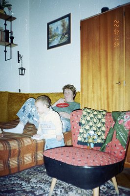 A young boy in denim overalls sits on a patterned sofa next to a woman holding a plate. The room features dated decor: a land...