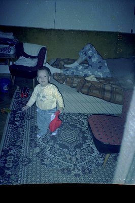 A young toddler stands on a patterned rug, holding a red toy. A woman is partially visible on a couch in the background, disp...