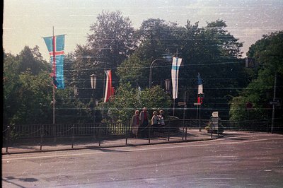 A group of people walk along a paved pedestrian walkway with metal railing, lined with vibrant flag banners. Lush greenery fi...