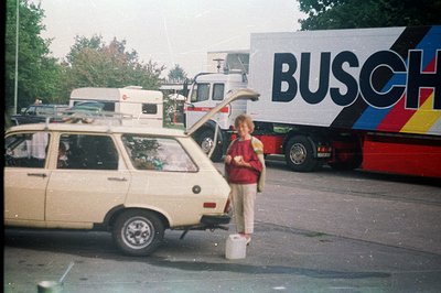 A young boy stands beside a beige Lada estate car, a bucket at his feet. A large "BUSCH" branded truck is visible in the back...