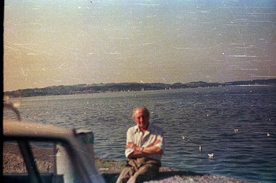 A seated older man, arms crossed, overlooks a wide body of water with distant hills. The photograph exhibits characteristics ...