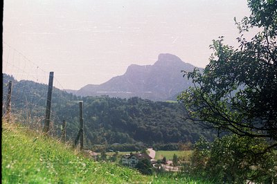 Hilly, forested landscape with a distinctive, jagged mountain peak dominating the view. A small village and reservoir are nes...
