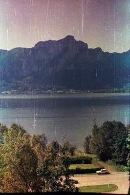 Lush foliage frames a scenic view of a lake reflecting imposing mountains. A vintage car is parked on a paved drive leading t...