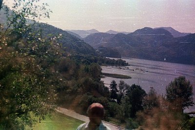 Scenic view of a river valley, likely the Hudson River, framed by lush, forested hills. A person stands at the edge of a lawn...