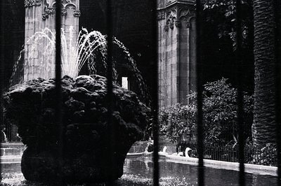 A tiered fountain sits within a courtyard, framed by wrought iron bars. Behind, a Gothic Revival-style building features poin...