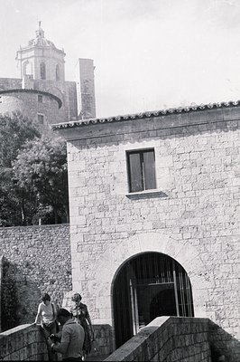 Stone building with arched gateway & partial view of a tower church. Three figures visible – two adults, one child – near the...