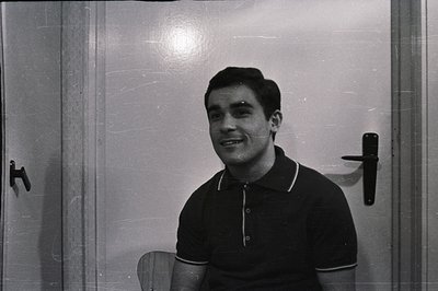 A smiling man in a black polo shirt sits near a door. The photograph’s minimalist setting highlights the subject’s casual sty...