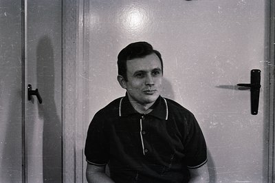 A monochrome portrait of a man seated against a plain wall and door. He wears a black polo shirt with white trim. Simple, geo...
