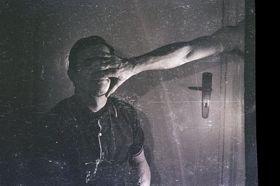 A man blocks his face with his hand in a grainy, high-contrast black and white image. Visible wall and door hardware suggest ...