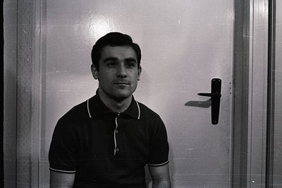 A portrait of a man in a black polo shirt stands against a door. The composition is simple; focus on the subject’s direct gaz...