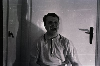 Man with an open mouth, appearing to shout or exclaim. Portrait-style, against a plain door background. He wears a short-slee...