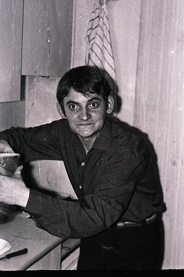 A candid black and white portrait of a man in a button-down shirt, holding a small bowl. The photograph features a grainy tex...
