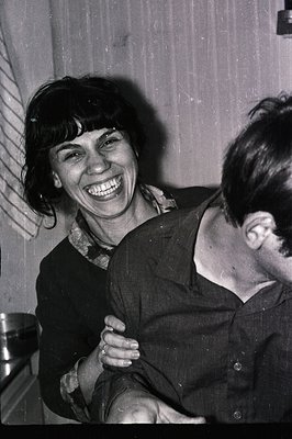 A close-up captures a smiling woman with dark, short hair and a patterned shirt, partially embracing a man whose face is crop...