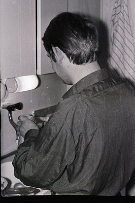 A young man, viewed from behind, meticulously works on a watch or clock mechanism in what appears to be a workshop or repair ...