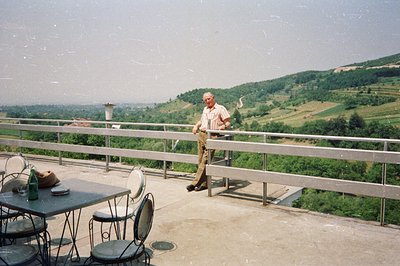 An older man stands near a metal railing overlooking a lush, green, hilly landscape. A table with drinks and cutlery sits nea...