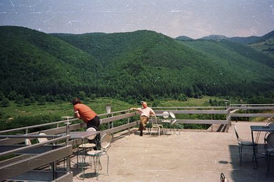 A rooftop terrace with two figures overlooking a lush, green, mountainous landscape. The architectural style suggests a 1970s...