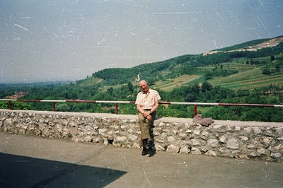 A senior gentleman, wearing a short-sleeved shirt & corduroy trousers, sits on a stone retaining wall overlooking a hillside ...