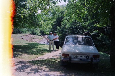 A vintage sedan, likely a Lada, sits on a dirt road flanked by dense woodland. Two figures, a man in a hat and a woman, stand...