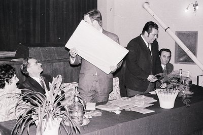 Formal meeting or presentation scene; three men and a woman seated behind a table. A large chart or display board is being pr...