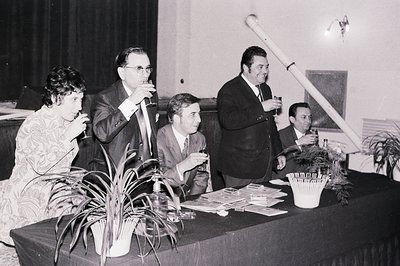 Formal event scene, c. 1960s-70s. Four individuals at a table, likely judging a competition—note cards visible. Man in glasse...