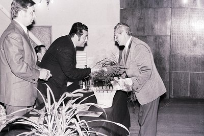 Three men in suits interact near a table covered with brochures and a decorative plant arrangement. A formal reception or awa...