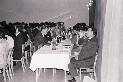 Formal dinner scene, likely a banquet or reception. Guests in suits and dresses seated around a long table covered with a whi...