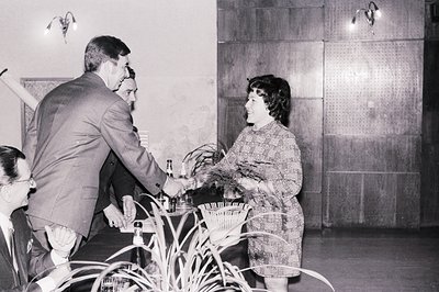 A man in a suit serves a drink to a woman in a patterned dress, likely at a social event. Interior setting features wood-pane...