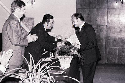A formal presentation; two men in suits offer what appears to be a ceremonial plaque or diploma to a third, who smiles and ge...
