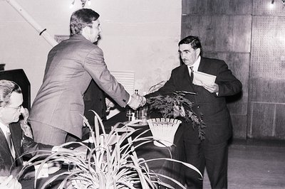 Two men share a formal handshake, one extending a document. The man on the right wears a dark suit and tie, while the other h...