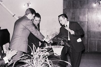 Three men in dark suits and slicked-back hair stand around a table covered with small bottles and a plant. Likely a formal ev...