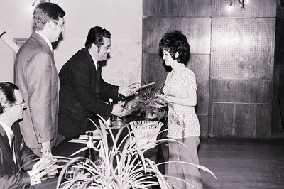 Formal portrait captures an award ceremony, likely in the 1960s. A man in a suit presents a plaque to a woman with a stylish ...