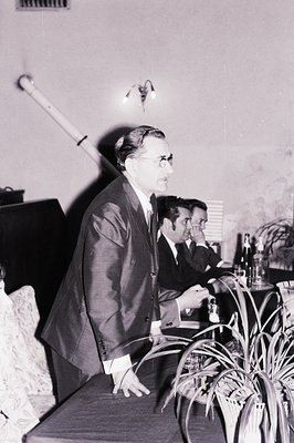 Man in a suit stands, appearing to address a seated group. A long, white object (possibly an instrument) dominates the left f...