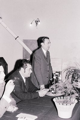 Men seated around a dark, formal table during an event. One man is smoking, another is speaking while gesturing. A potted pal...