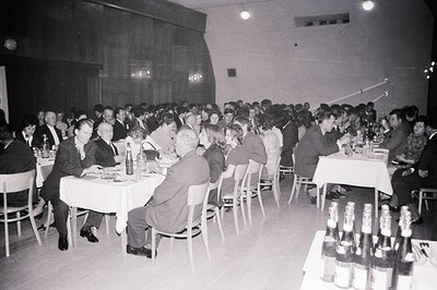 Large group seated at tables covered with white cloths in a hall. Men in suits and ties, some women in dresses. Bottles of wi...