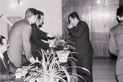 Formal gathering, circa 1960s. Three men in suits observe another man seated, appearing to present a small trophy or award. I...