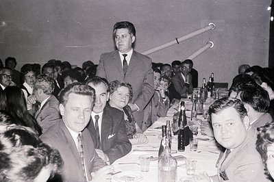 A formal dinner or banquet scene, likely 1960s. Guests seated at tables adorned with dark bottles & glassware. Men in suits, ...