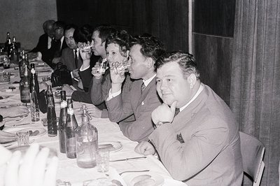 Group scene at a formal dinner. Several men and women in suits and dresses raise glasses in a toast. A long table covered in ...