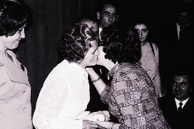 A formal, intimate moment captured: a woman in a white, lace-trimmed dress receives a kiss on the cheek from another woman wi...