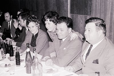 A candid group portrait at a formal dinner. Four men and two women are seated at a long table, enjoying a meal with drinks an...