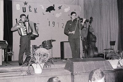 Three musicians perform onstage, featuring an accordionist, bassist, and drummer. A backdrop displays "BUEK" and "1972." Stag...