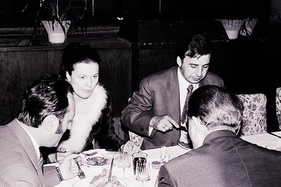 Four formally dressed individuals seated around a table, likely dining in a restaurant. A woman with a fur collar is central,...