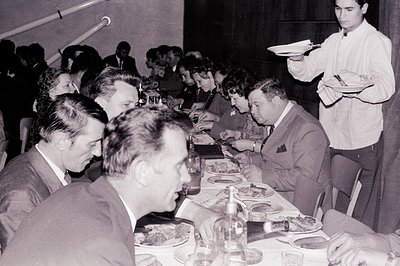 A group of formally dressed patrons dine at a long table during what appears to be a banquet or formal event. A server carrie...