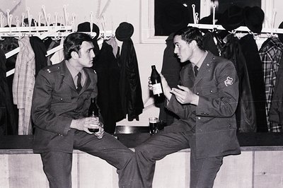 Two uniformed men, likely military personnel, are seated and holding bottles, perhaps of beer or wine. Background shows a sto...
